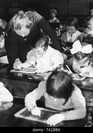 La classe di un GIR'ls classe siedono nei banchi di scuola e di scrivere con il gesso sulle loro schede di scrittura. Un insegnante è aiutare una delle ragazze. Foto Stock