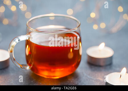 Tè nero in tazza di vetro con candele e ghirlanda luminosa. Sfondo grigio. Copia dello spazio. Foto Stock