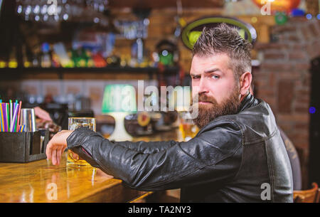 Brutale parigamba uomo barbuto sedersi al banco bar bere birra. Ordine di bere alcol. Il bar è un luogo rilassante hanno bere e rilassarsi. Uomo con barba trascorrere il tempo libero nella barra di colore scuro. Hipster rilassante presso il bar con birra. Foto Stock