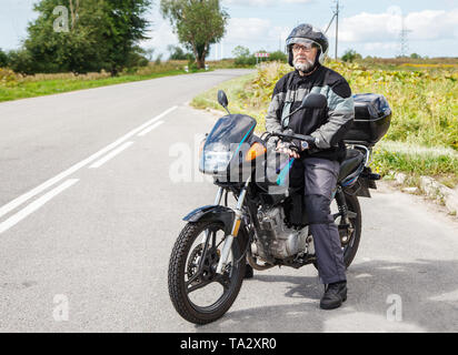 Anziani motociclista che indossa una giacca e occhiali con un casco seduto su una motocicletta sulla strada aperta su soleggiate giornate estive Foto Stock