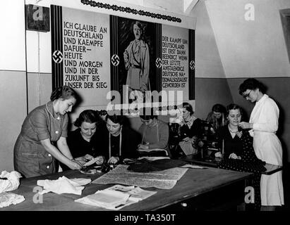 Foto dei partecipanti di un corso di cucitura del Reichsmuetterdienst. Sulla sinistra, il lavoratore sociale che guida le altre donne. Sullo sfondo di una propaganda poster con Adolf Hitler e la scritta "Io credo in Germania e lottare per essa - di oggi e di domani e in futuro fino a quando non si vince. La lotta per la Germania è finita solo quando l'ultima tedesco decente è diventato un socialista nazionale'. Foto di Propaganda Company (PK) Foto Stock