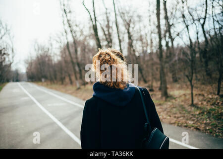 Vista posteriore della giovane donna vestita di nero coat a piedi giù per la strada. Una ragazza con una coda di cavallo acconciatura va lungo il parco. Foto Stock