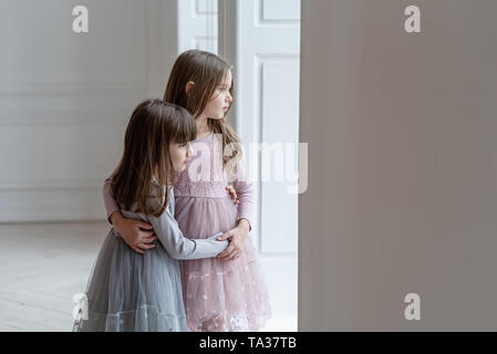 Due ragazze commovente abbraccio e guardare fuori dalla finestra. Gravi piccole sorelle in belle abiti stare vicino a una finestra. Foto Stock