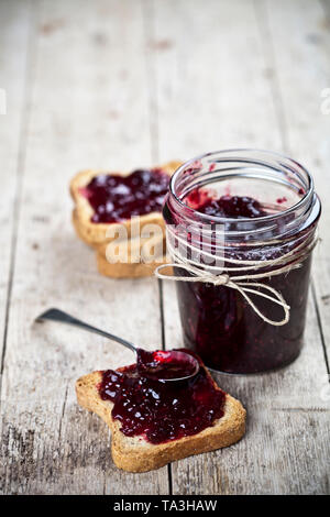 Cereali tostati le fette di pane e jar con fatti in casa frutti di bosco Confettura e closeup cucchiaio su tavola in legno rustico sfondo. Sweet Food per la prima colazione con Foto Stock