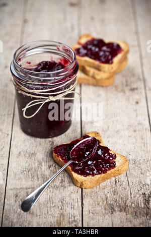 Cereali tostati le fette di pane e jar con fatti in casa frutti di bosco Confettura e closeup cucchiaio su tavola in legno rustico sfondo. Sweet Food per la prima colazione con Foto Stock