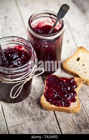 Cereali tostati le fette di pane e di vasi fatti in casa con frutti di bosco e confettura di ciliegie e cucchiai closeup su tavola in legno rustico sfondo. Sweet Food per b Foto Stock