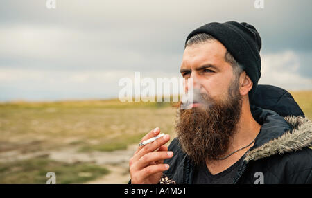 Uomo Barbuto il fumo di sigarette, bello hipster ritratto con faccia grave ha una lunga barba in hat outdoor su sfondo naturale Foto Stock