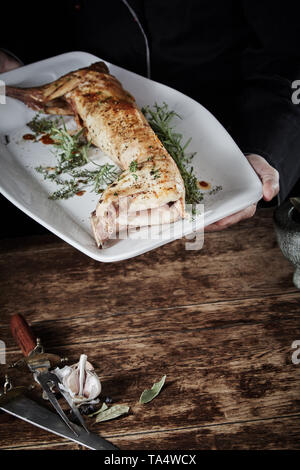 Arrosto di capriolo di coniglio selvatico su un grande piatto bianco su un tavolo rustico con in primo piano lo spazio di copia con aglio fresco ed erbe aromatiche nell'angolo Foto Stock