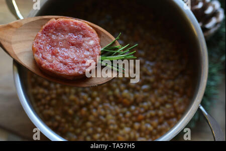 Cenone di Capodanno in stile italiano. Vista dall'alto di una fetta di cotechino e rosmarino sul cucchiaio. Sfondo con la pentola con le lenticchie. Foto Stock