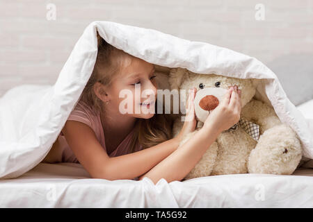 Adorabile bambina ragazza che gioca con Teddy bear nel letto nella mattina. bambino con un orsacchiotto sotto i coperchi Foto Stock