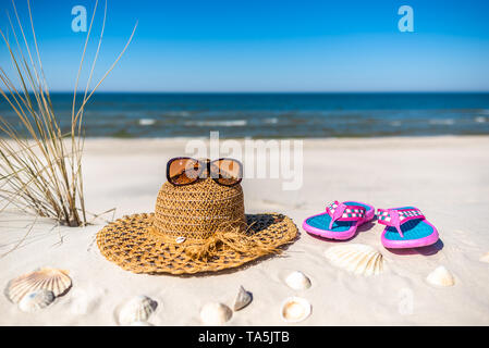 Sfondo con vacanza estate spiaggia accessori oltre il Mar Baltico con sabbia bianca e blu del cielo. Bicchieri, cappello per il sole e flip flop. Foto Stock