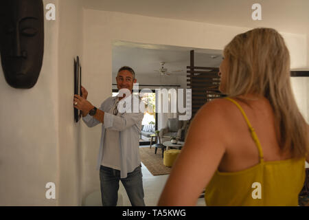 Uomo maturo appeso cornice alla parete mentre donna interagendo con lui Foto Stock