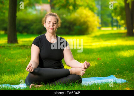 Flessibile e concentrata di donna è sovradimensionata meditando sul prato nel parco nella posizione del loto Foto Stock