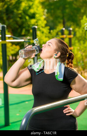 Dimensioni plus ragazza beve acqua pulita da un flacone dopo le attività sportive nel Parco Foto Stock