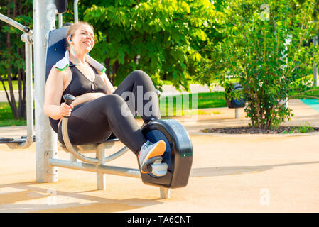 Felice e attiva donna sovradimensionato facendo esercizio su una bicicletta stazionaria in un parco della città Foto Stock