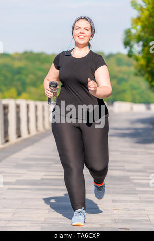 Taglie donna con una bottiglia di acqua durante la sua Morning jog in un parco della città Foto Stock