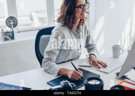 Femmina giovane fotografo lavorando sul computer utilizzando il tampone di disegno a scrivania in ufficio. Giovane donna caucasica tramite digital tavoletta grafica e disegno a penna Foto Stock