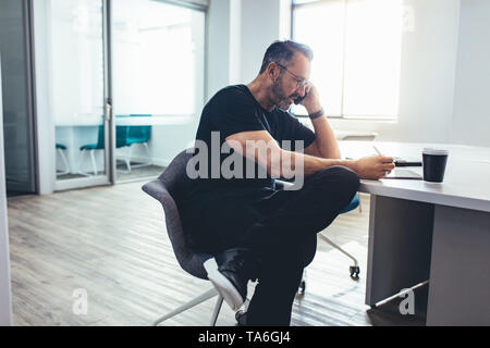 Imprenditore maschio guardando la sua tavoletta digitale e parlando al cellulare mentre è seduto in ufficio. Imprenditore occupato a lavorare in ufficio. Foto Stock