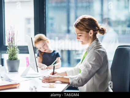 Imprenditrice con figlia piccola seduta in un ufficio, lavorando. Foto Stock