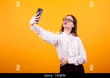 Ragazza giovane e carina facendo una selfie e facendo le labbra di anatra alla fotocamera in studio su sfondo giallo. Bella ragazza giovane isolato. Foto Stock