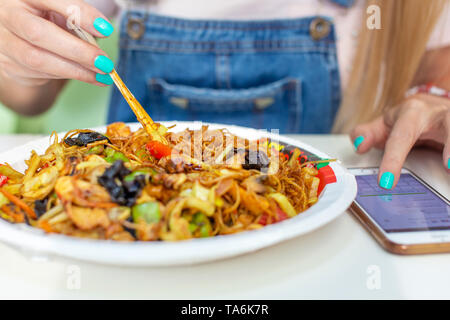 La donna cinese di mangiare a base di noodle con pollo e messaggistica sullo smartphone Foto Stock