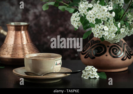 Close-up di una tazza di caffè e un ramo di fioritura spirea in un vaso di ceramica su sfondo scuro Foto Stock