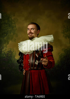 Giovane uomo come un cavaliere medievale su studio scuro dello sfondo. Ritratto in chiave di basso del modello maschile in costume retrò. Puntamento e sorridente. Le emozioni umane, co Foto Stock