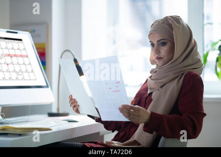 Dark-eyed insegnante indossa hijab seduto vicino a un computer Foto Stock