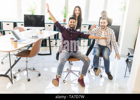Carefree eccitato diversi lavoratori aventi fun riding oh sedie celebrando venerdi insieme, felici i dipendenti godono di concorrenza divertenti ridere insieme fe Foto Stock