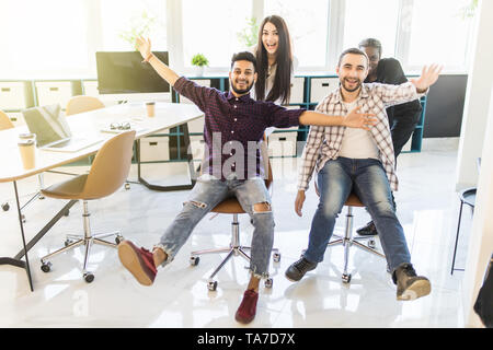 Avvio multiethnics team di sviluppatori di software per divertirsi durante la corsa su sedie da ufficio,entusiasti diversi dipendenti ridere godendo di attività divertenti a Foto Stock