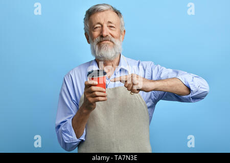 Felice vecchio uomo in piedi con la tazza monouso di caffè e che puntano ad esso isolato su sfondo blu. Buona scelta.il caffè migliore per le persone anziane Foto Stock