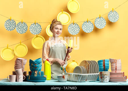 Sorridente giovane bella casalinga in grigio grembiule spugna di tenuta isolata su sfondo giallo.job, professione, professione, Foto Stock