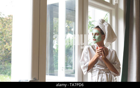 Donna in accappatoio indossando maschera facciale, guardando attraverso la finestra Foto Stock
