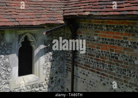 Rotto grondaia in metallo che pendono dal rosso tetto di tegole contro una chiesa parete di pietra focaia con piccola finestra sulla sinistra Foto Stock