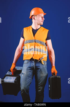 Professional repairman concetto. L uomo nel casco, hard hat detiene il toolbox e valigia con utensili, sfondo blu. Tuttofare, aggiustatore sulla faccia concentrato porta sacchetti pesanti con attrezzature professionali. Foto Stock