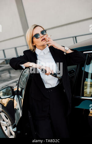 Attraente donna bionda in occhiali da sole il fumo di sigarette e tenendo la pistola vicino auto Foto Stock