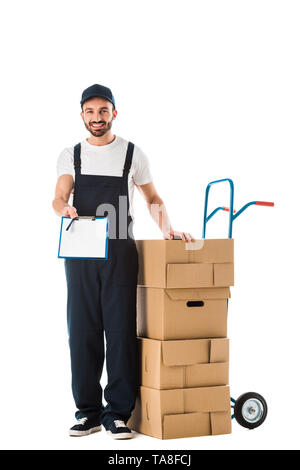 Consegna sorridente uomo azienda appunti con schermo vuoto mentre sta in piedi vicino a mano carrello caricato con scatole di cartone isolato su bianco Foto Stock