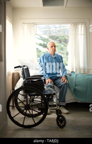 Senior contemplativa uomo seduto sul bordo del suo letto accanto a una sedia a rotelle. Foto Stock
