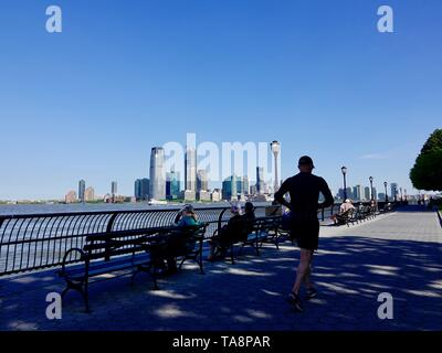 Pareggiatore passando da persone rilassante sulle panchine lungo il fiume Hudson, Batteria Esplanade, New York, NY, STATI UNITI D'AMERICA. Foto Stock