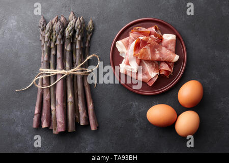 Mazzetto di freschi Asparagi viola, con uova e bacon in piastre su arrugginito sfondo testurizzata Foto Stock