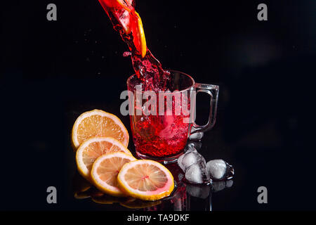 Estate bevanda ghiacciata - hibiscus tè freddo con ghiaccio, limone e menta. Hibiscus tè freddo versando in bicchiere con ghiaccio isolato riflettente sulla superficie nera. Hi Foto Stock