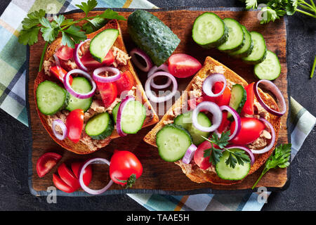 Aprire panini con fegato di merluzzo pate, cetriolo, pomodoro e cipolla rossa anelli su di un tagliere con ingredienti su di una tavola di cemento, vista da sopra, Foto Stock