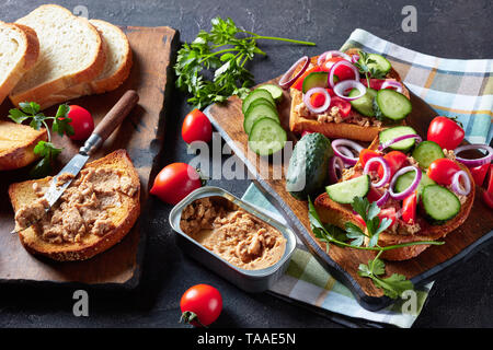 Close-up di toast con fegato di merluzzo pate, guarnita con fette di cetriolo, pomodoro e cipolla rossa anelli su di un tagliere con ingredienti su una scheda da cucina Foto Stock