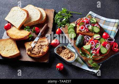 Close-up di toast con fegato di merluzzo pate, guarnita con fette di cetriolo, pomodoro e cipolla rossa anelli su di un tagliere con ingredienti su una scheda da cucina Foto Stock