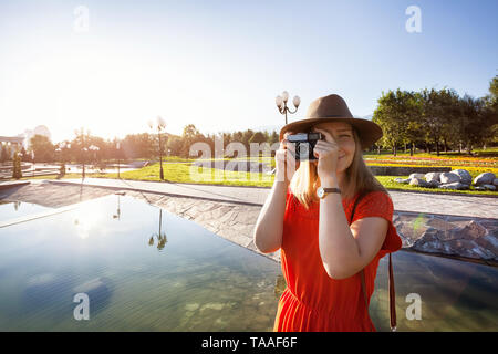 Felice donna turistica nel cappello e abito arancione con fotocamera vintage nel parco di sunrise Foto Stock