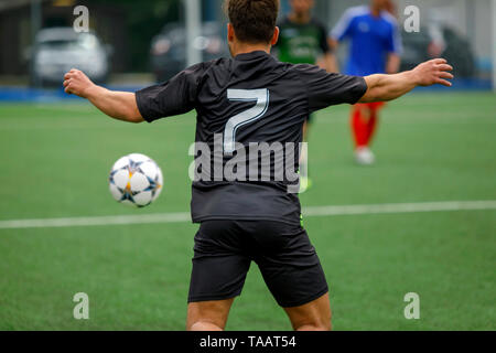 Giocatore di calcio in un campo di calcio insegue la palla in un gioco d'azione. Un Uomo in camicia nera e pantaloncini, incorniciato da dietro nel verde del campo di calcio. Foto Stock