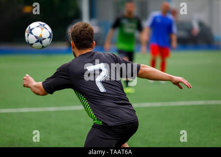 Giocatore di calcio in un campo di calcio insegue la palla in un gioco d'azione. Un Uomo in camicia nera e pantaloncini, incorniciato da dietro nel verde del campo di calcio. Foto Stock