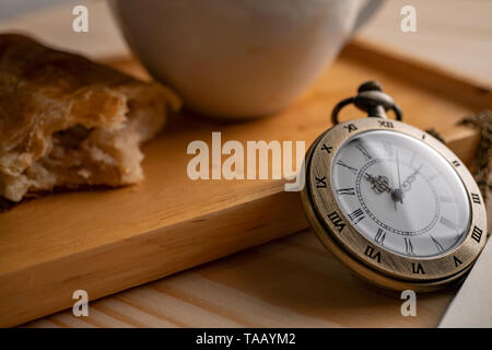 Oro antico orologio da tasca posta sul lato del vassoio in legno con un bianco tazza da caffè e la torta che è stato picchiato Foto Stock