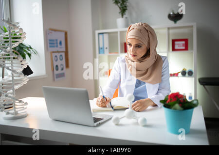 Utilizzando laptop. Scienziato medico indossa velo a lavorare utilizzando laptop e rendendo alcune note Foto Stock