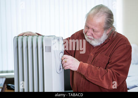 Senior uomo regola la temperatura di olio elettrico riscaldatore in ufficio nel periodo invernale Foto Stock